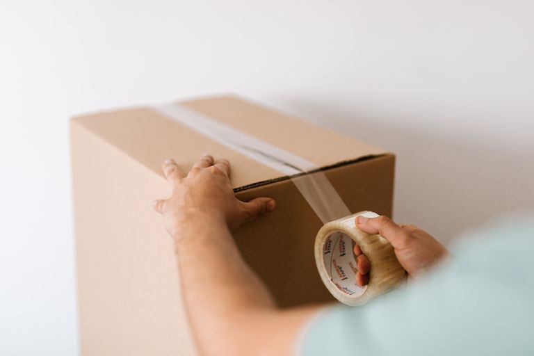 Crop faceless male sealing carton container with belongings by using tape against white plain wall before moving out